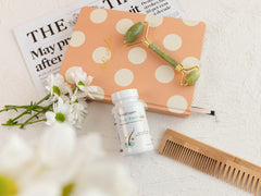 SanaExpert Haar Forte Gold Flasche vor einem Kosmetikbeutel mit Polka-Dots, einer Gesichtsroller und frischen Blumen, neben Zeitschriften und einem Holzkamm.