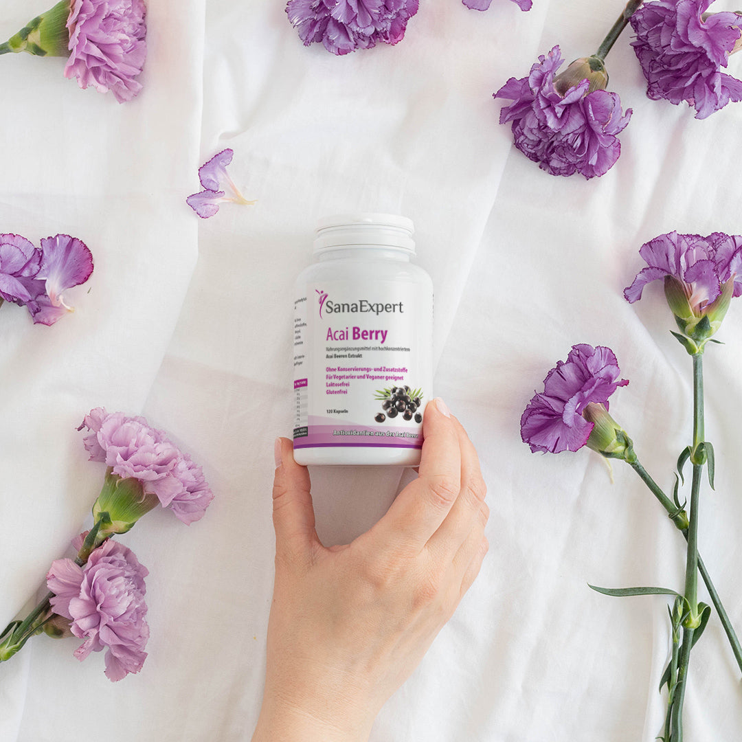 Hand hält eine Flasche SanaExpert Acai Berry Nahrungsergänzungsmittel, umgeben von violetten Blumen auf weißem Stoffhintergrund